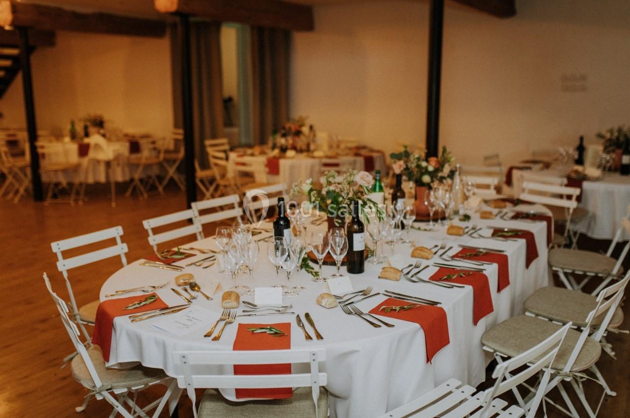 Salle de réception avec tables rondes dressées, nappes blanches, chemins de table rouges et décorations florales.