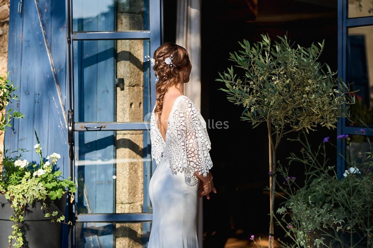 Une femme en robe de mariée blanche se tient devant une porte vitrée ouverte, entourée de plantes en pot.