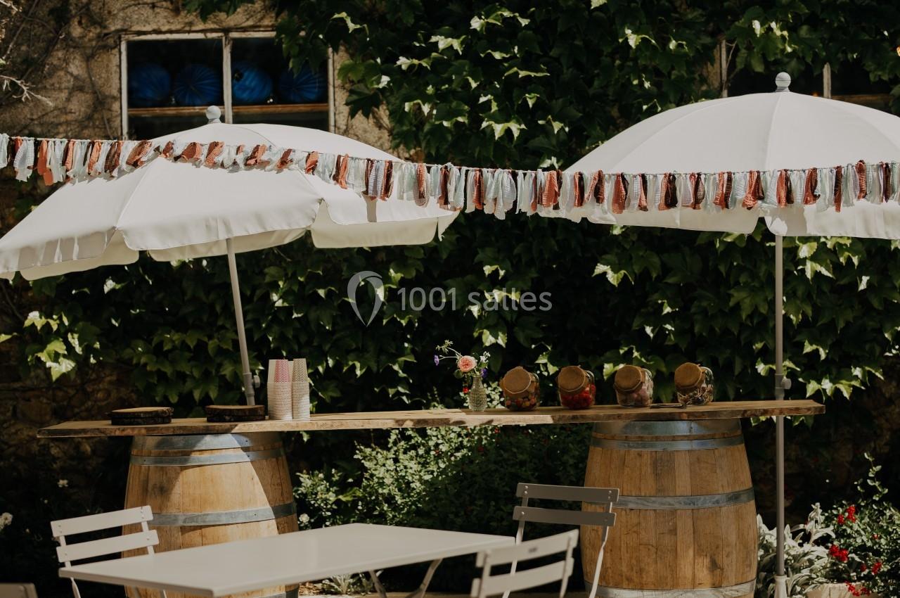 Table en bois posée sur des tonneaux, décorée de pots et guirlandes, sous des parasols blancs dans un jardin verdoyant.