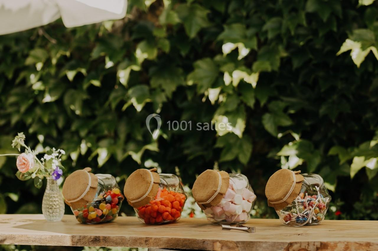 Quatre bocaux de bonbons colorés posés sur une table en bois devant un mur végétal et des parasols blancs.