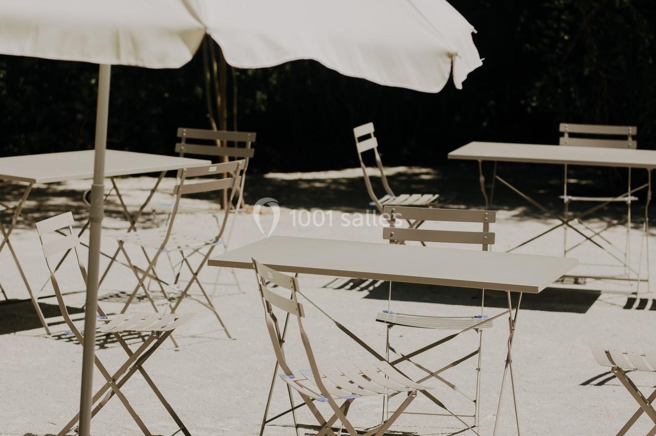 Chaises et tables en métal disposées sous un parasol blanc dans une cour ensoleillée avec des fleurs en arrière-plan.