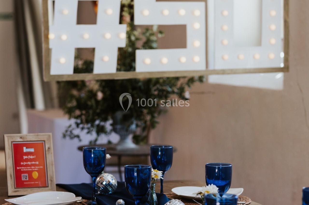 Table décorée avec vaisselle blanche, verres bleus et centre de table sombre, sous un panneau lumineux ’#50’.