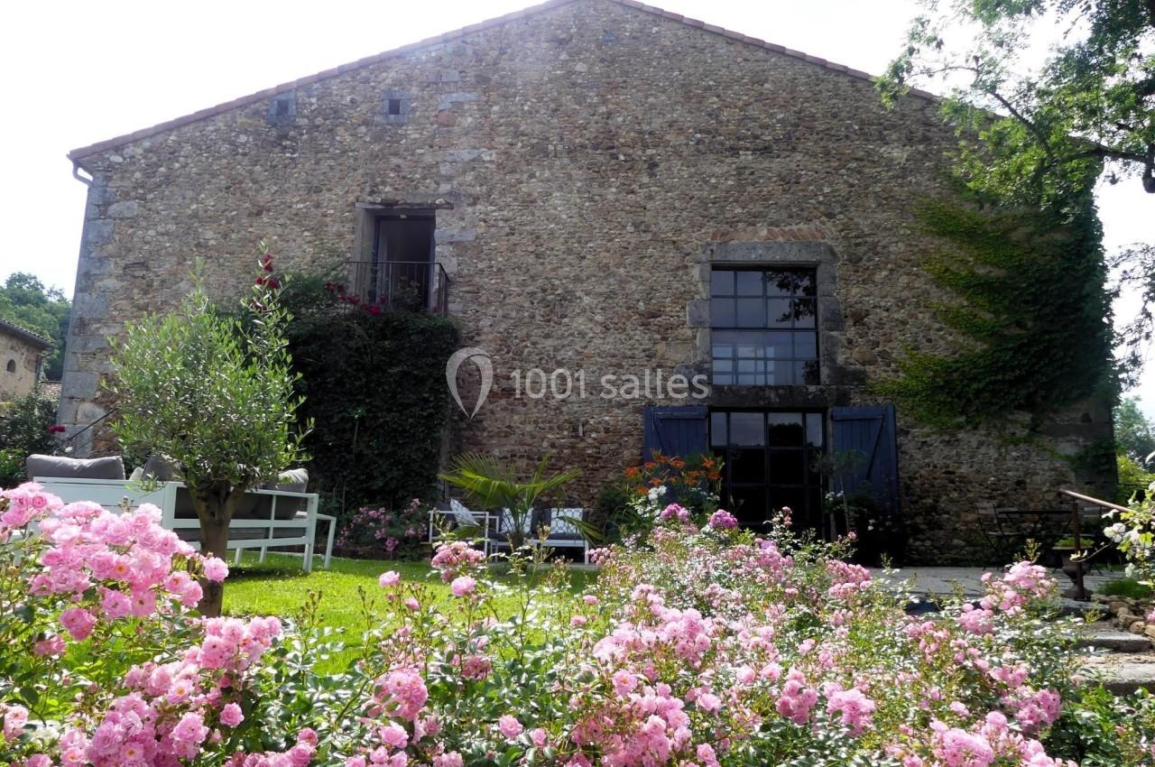 Façade en pierre d'une maison ancienne entourée de fleurs roses et de verdure, avec des fenêtres et des volets bleus.