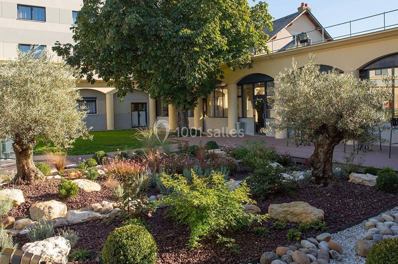 Jardin paysager avec arbres, arbustes et rocailles devant un bâtiment moderne à arcades.