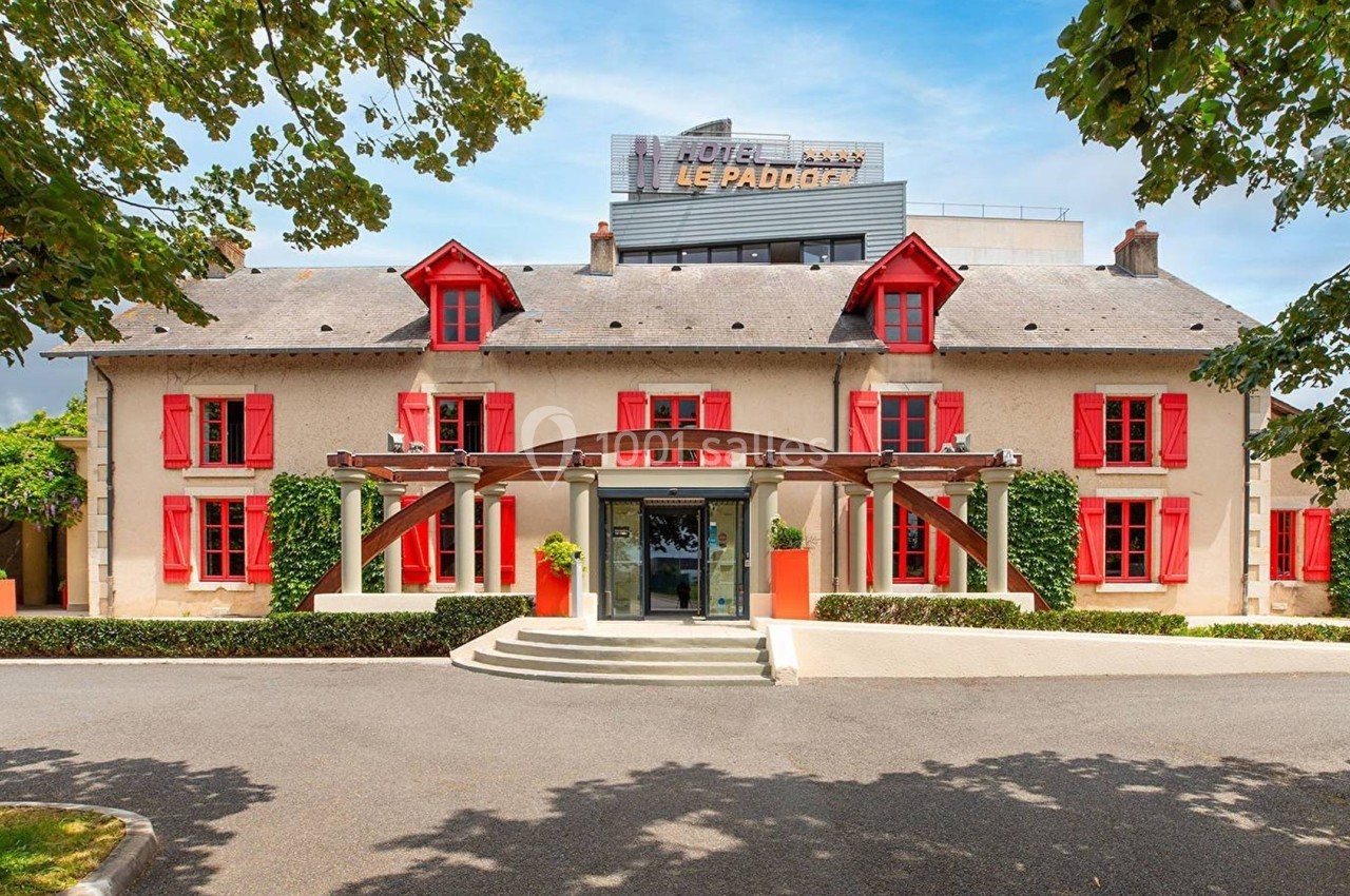 Façade d'un hôtel avec volets rouges, entrée centrale et enseigne visible sur le toit, entouré d'arbres.