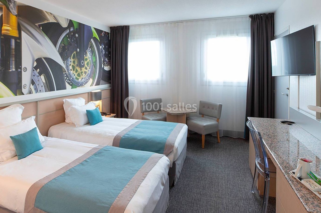 Chambre d'hôtel lumineuse avec deux lits simples, un bureau, une télévision murale et une décoration murale colorée.