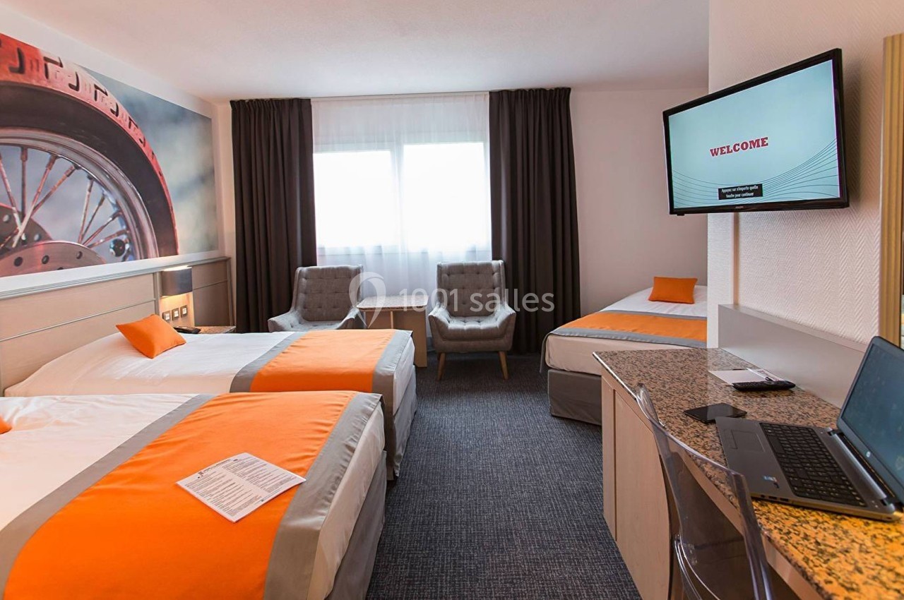 Chambre d'hôtel lumineuse avec trois lits, bureau, télévision, fauteuils, et décoration murale sur le thème d'une roue.