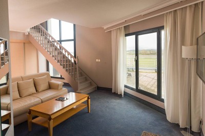 Salon lumineux avec canapé beige, table basse en bois, escalier, grande baie vitrée et vue sur un paysage extérieur.