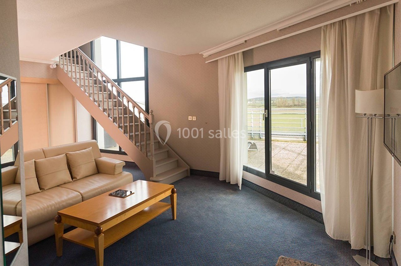 Salon lumineux avec canapé beige, table basse en bois, escalier, grande baie vitrée et vue sur un paysage extérieur.