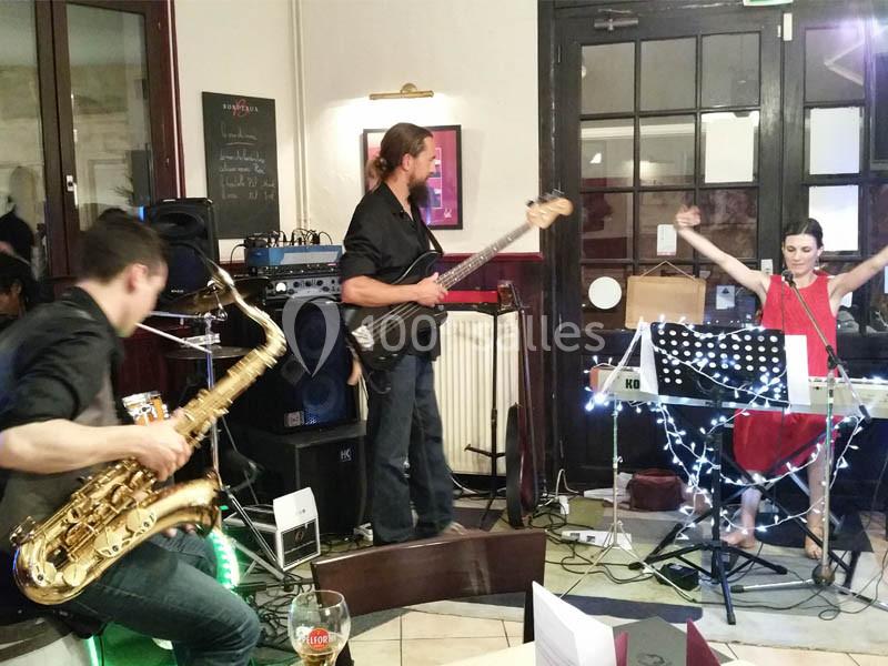 Un groupe de musiciens joue en intérieur, avec un saxophoniste, un bassiste et une claviériste chantant.