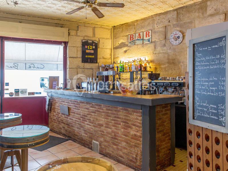 Intérieur d'un café avec un comptoir en briques, des bouteilles alignées, un tableau de menu et des tables rondes en bois.