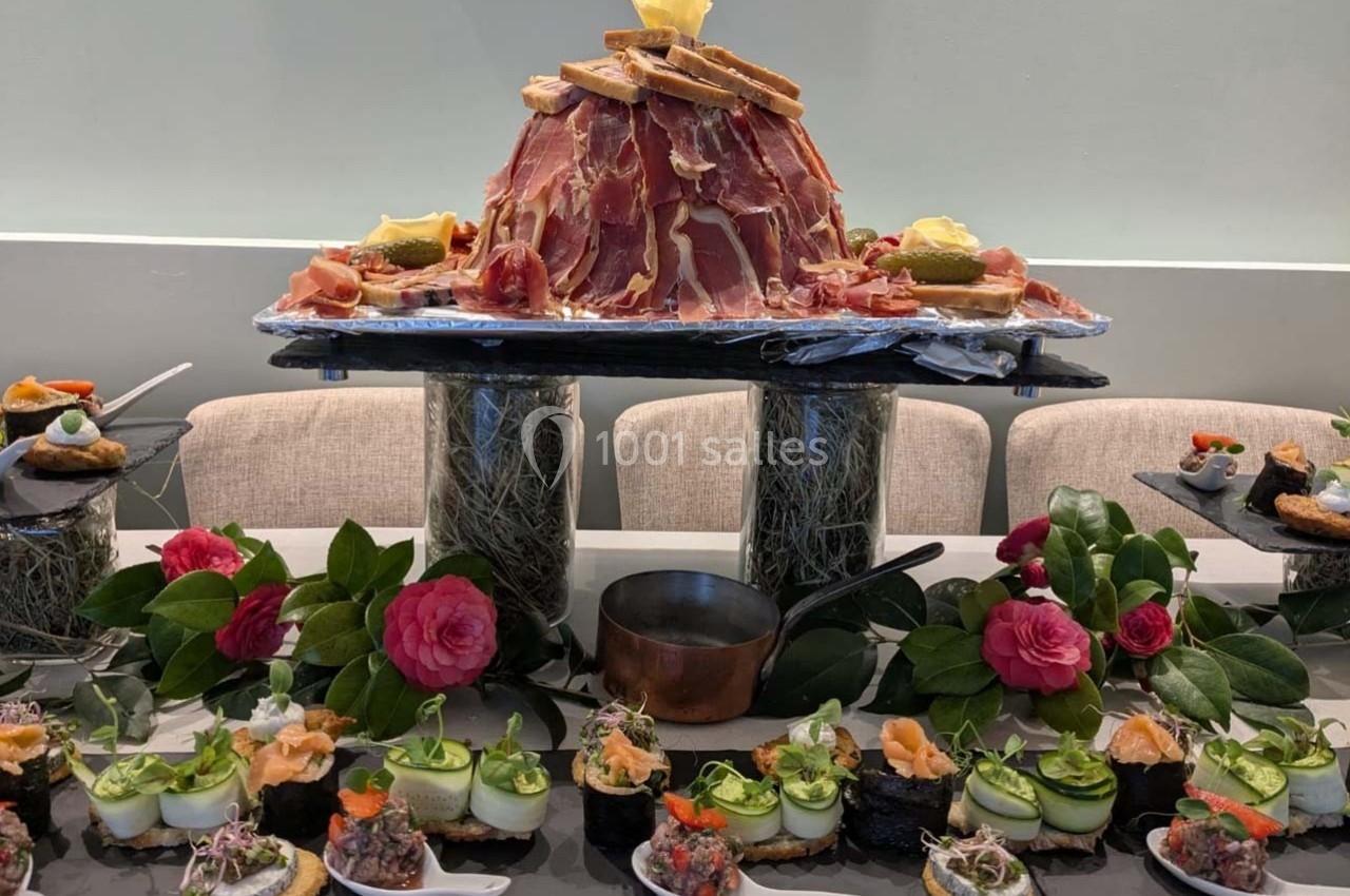Plateau de charcuterie en forme de montagne entouré de bouchées variées, décoré de fleurs et de verdure.