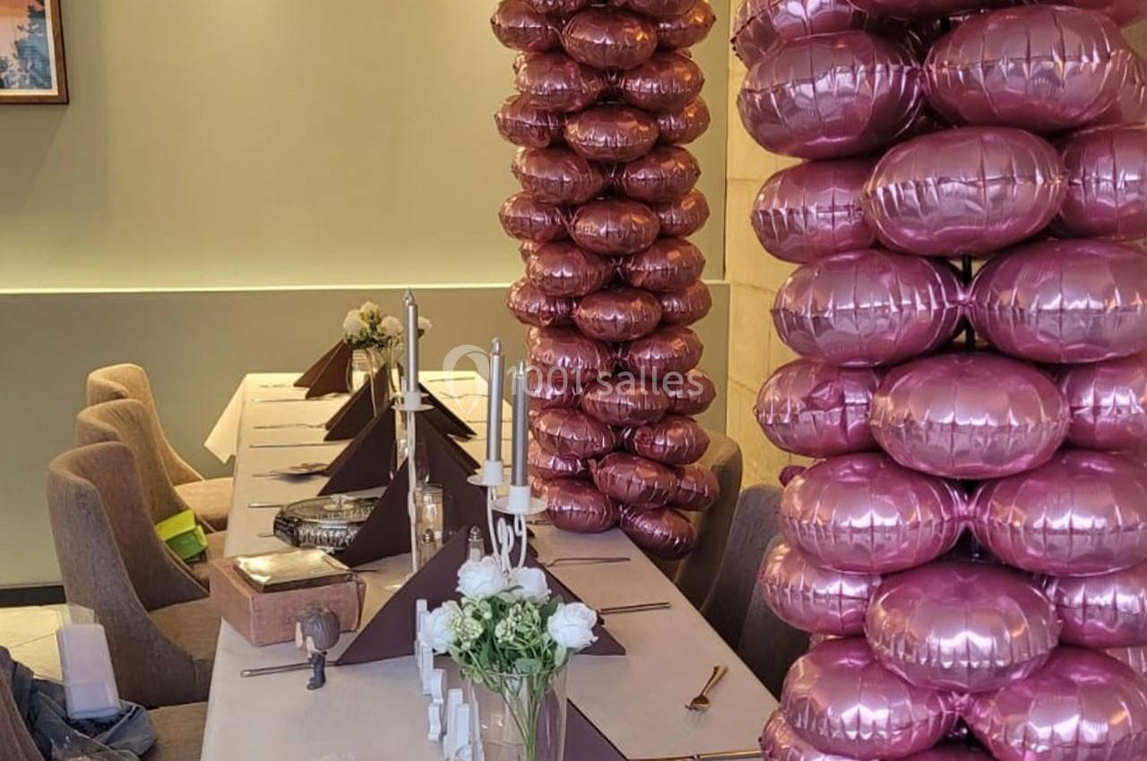 Table décorée avec des bougies, des fleurs blanches et des colonnes de ballons roses dans une salle élégante.