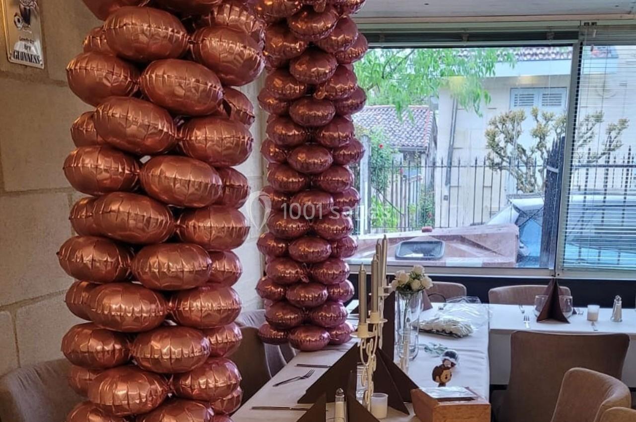 Deux colonnes de ballons roses métallisés décorent une salle avec des tables dressées près d'une baie vitrée.
