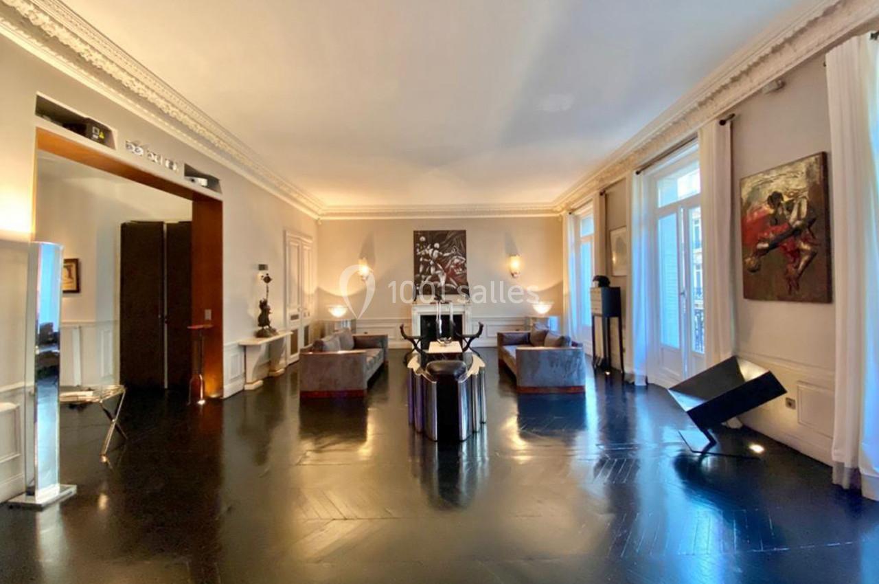 Salon spacieux avec parquet sombre, mobilier moderne, œuvres d'art aux murs et grandes fenêtres laissant entrer la lumière.