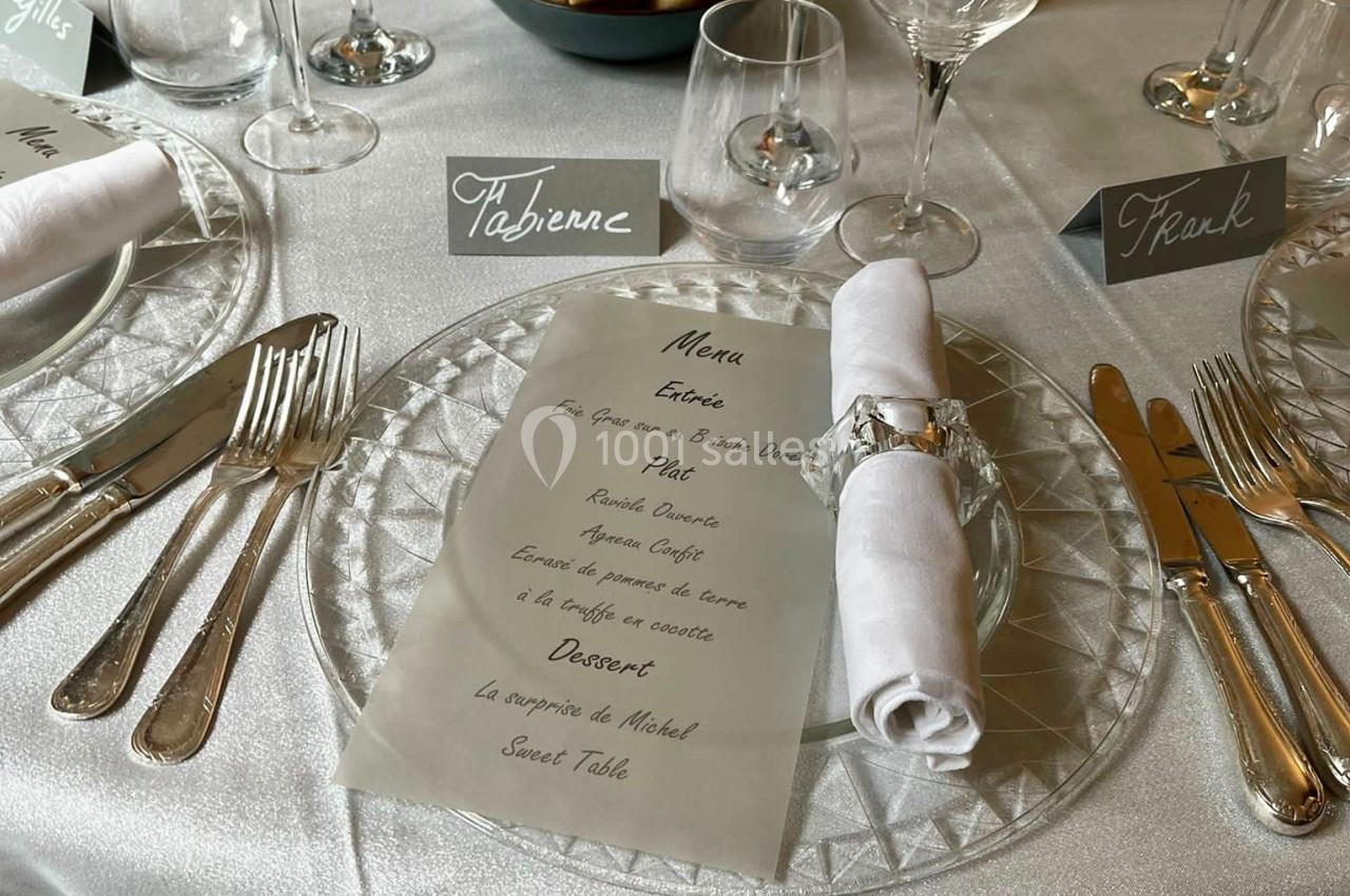 Table élégamment dressée avec menu, couverts, serviette roulée, verres et paniers de pain en arrière-plan.