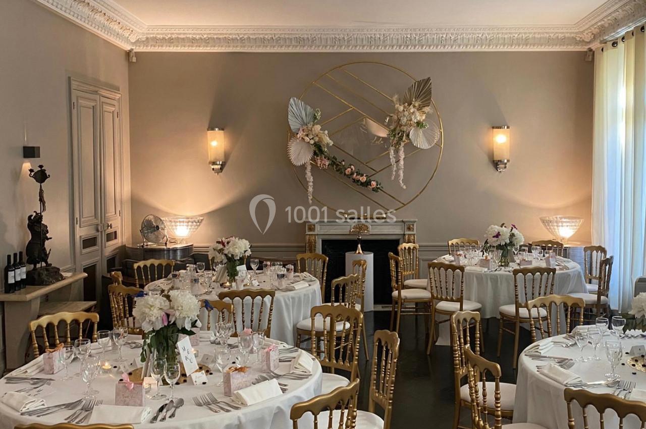 Salle élégante décorée pour un événement, avec tables rondes dressées, chaises dorées et décor floral mural.