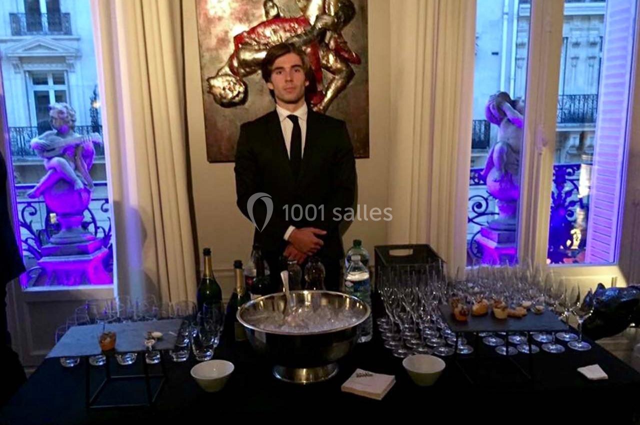 Un serveur en costume se tient derrière une table dressée avec des verres, bouteilles et amuse-bouches dans un intérieur…