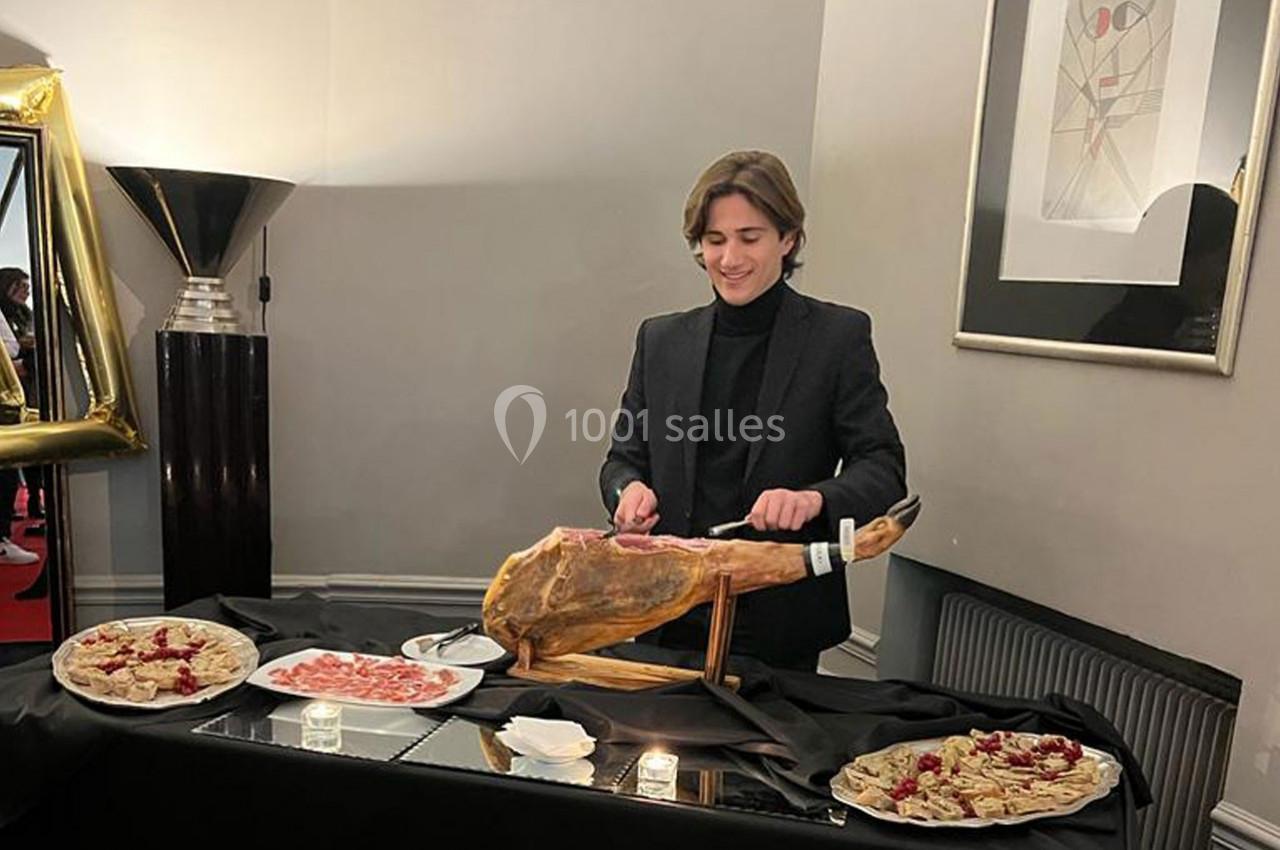 Un homme en costume tranche un jambon entier sur une table décorée avec des assiettes de charcuterie.