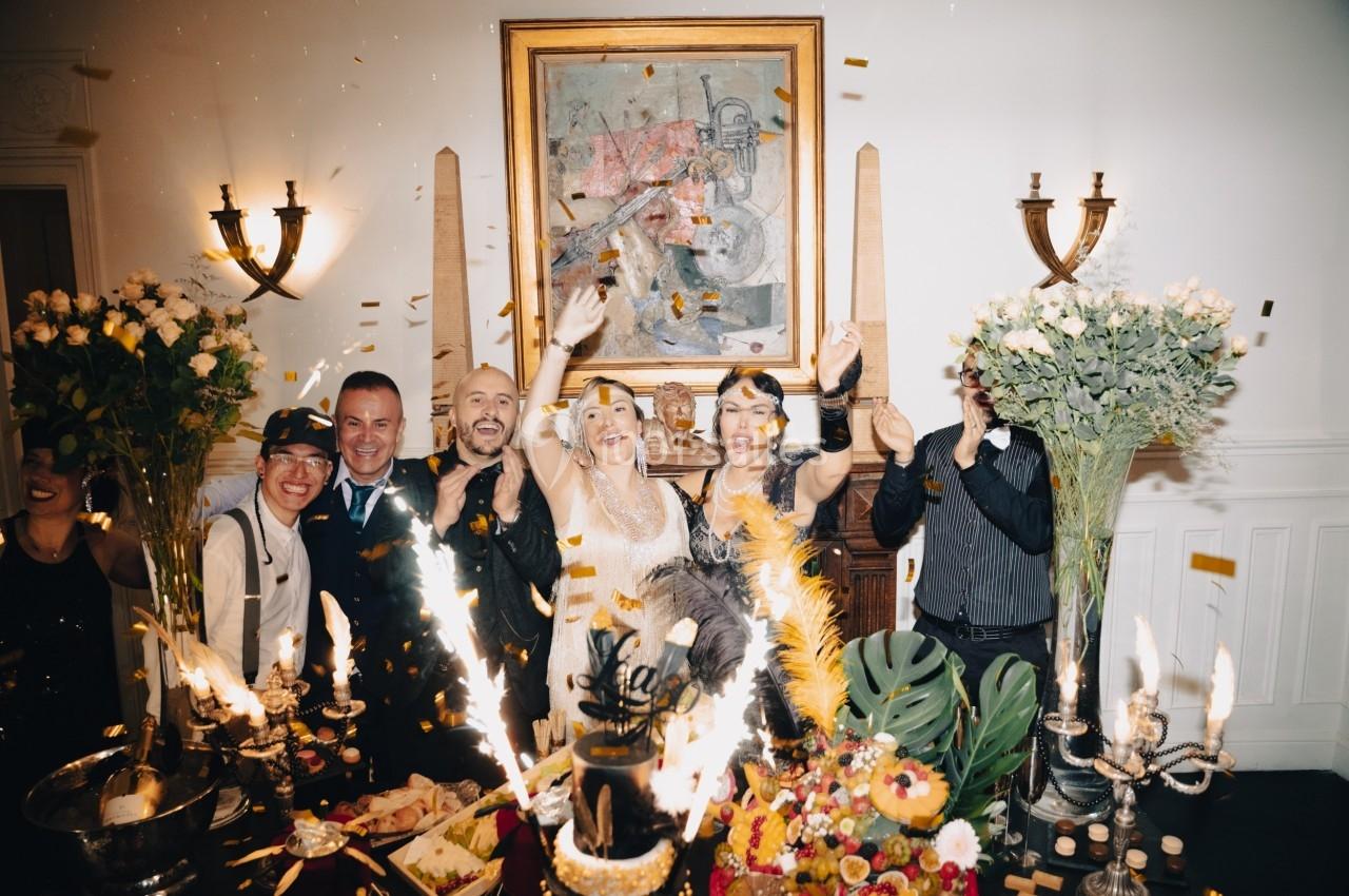 Groupe de personnes célébrant dans une ambiance festive avec confettis, décorations et table garnie de mets et bougies.