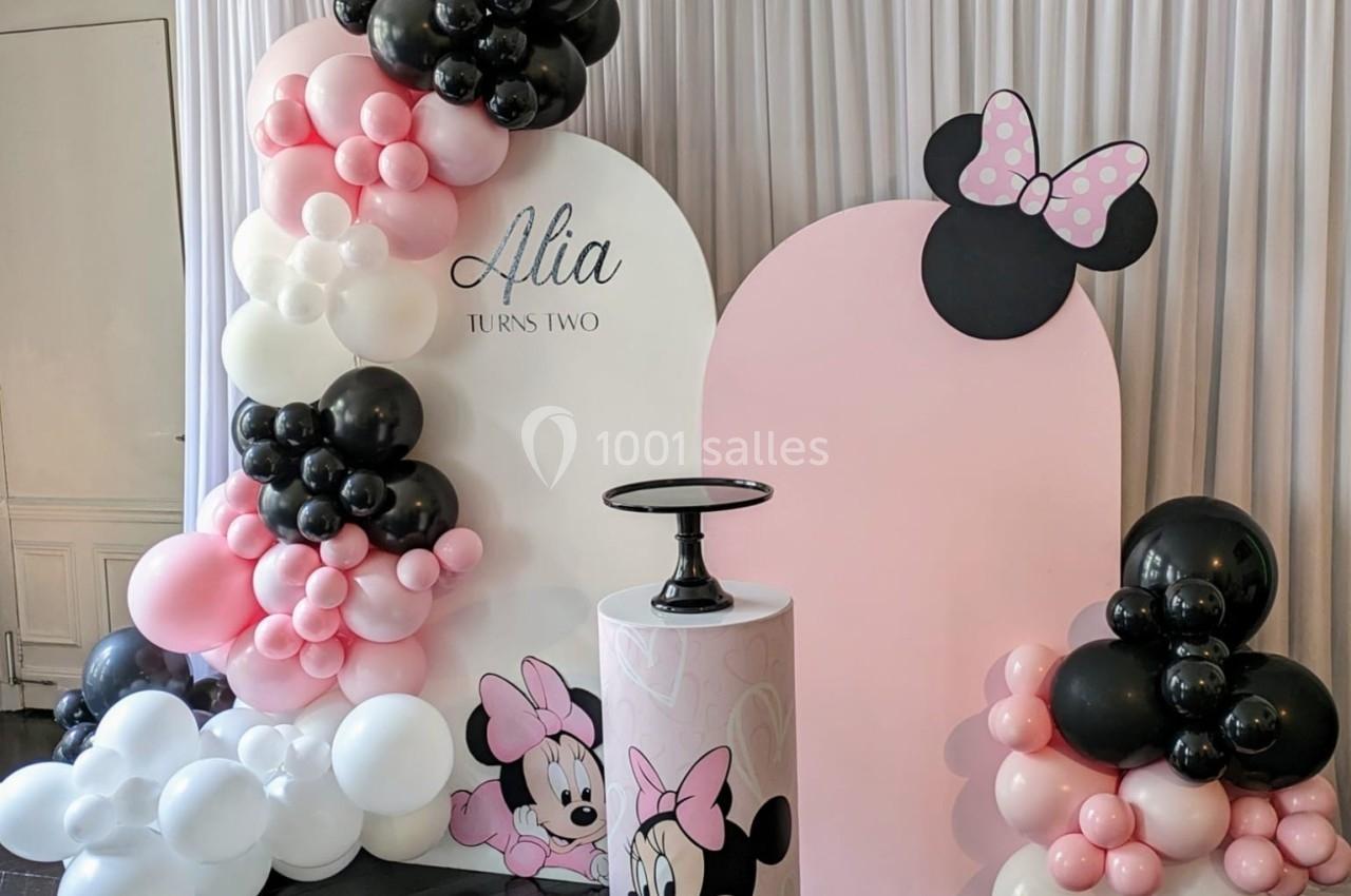 Décor festif avec ballons roses, blancs et noirs, et illustrations de Minnie Mouse devant un rideau blanc.