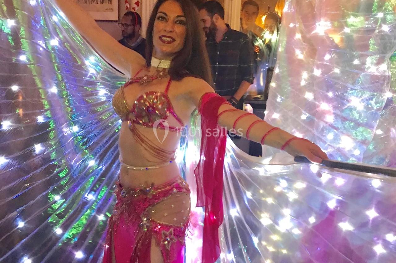 Danseuse en costume rose et ailes lumineuses, exécutant une performance dans une pièce éclairée.