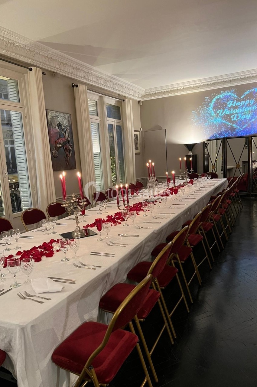 Grande table rectangulaire décorée pour un dîner, avec nappes blanches, chandeliers, pétales rouges et chaises rouges.