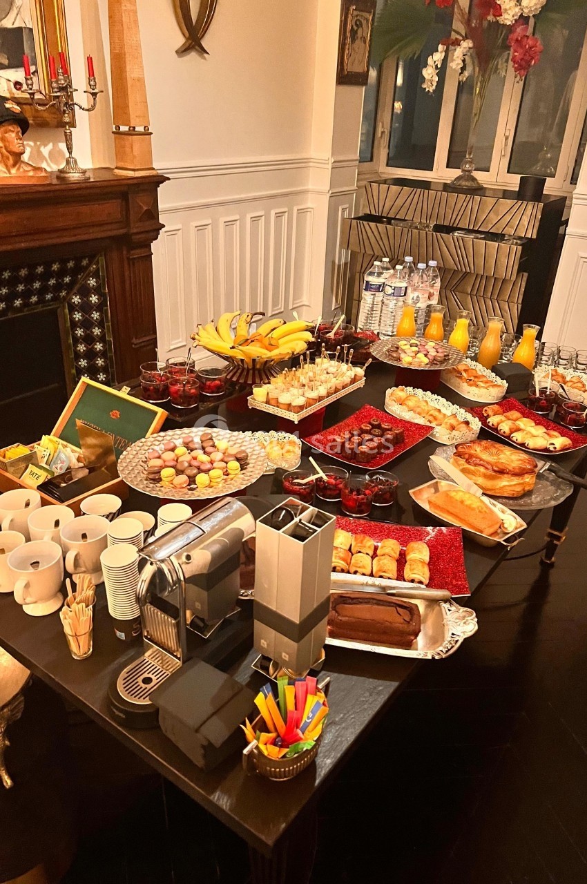 Table garnie de boissons, viennoiseries, fruits et desserts dans un intérieur chaleureux avec cheminée et décorations.