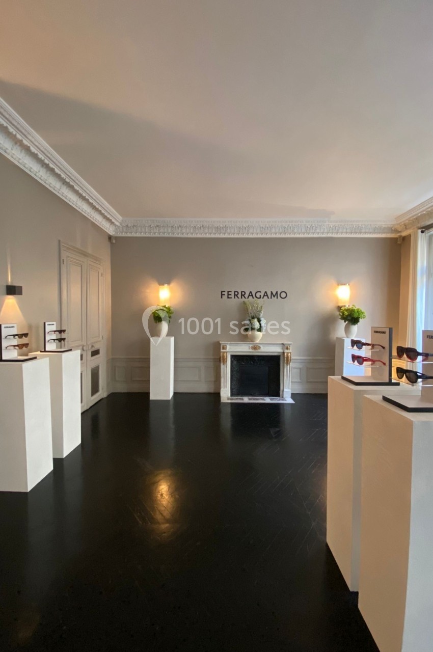 Salle d'exposition lumineuse avec des lunettes présentées sur des socles blancs et un mur marqué ’Ferragamo’.