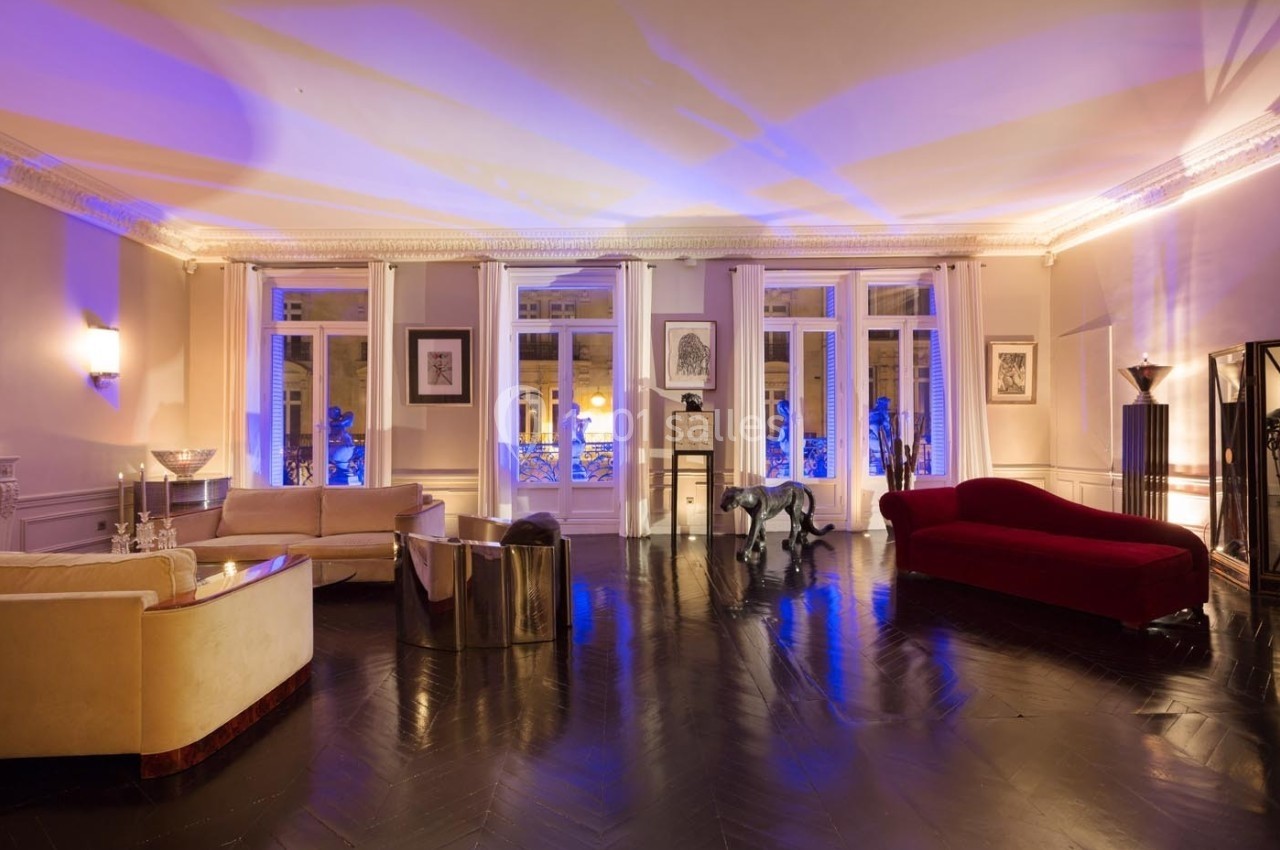 Salon spacieux avec parquet sombre, canapés, fauteuils design et éclairage tamisé, donnant sur de grandes fenêtres.