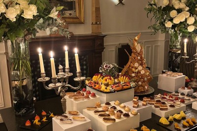Table garnie de pâtisseries variées, éclairée par des chandeliers, avec des fleurs et une pièce montée en arrière-plan.