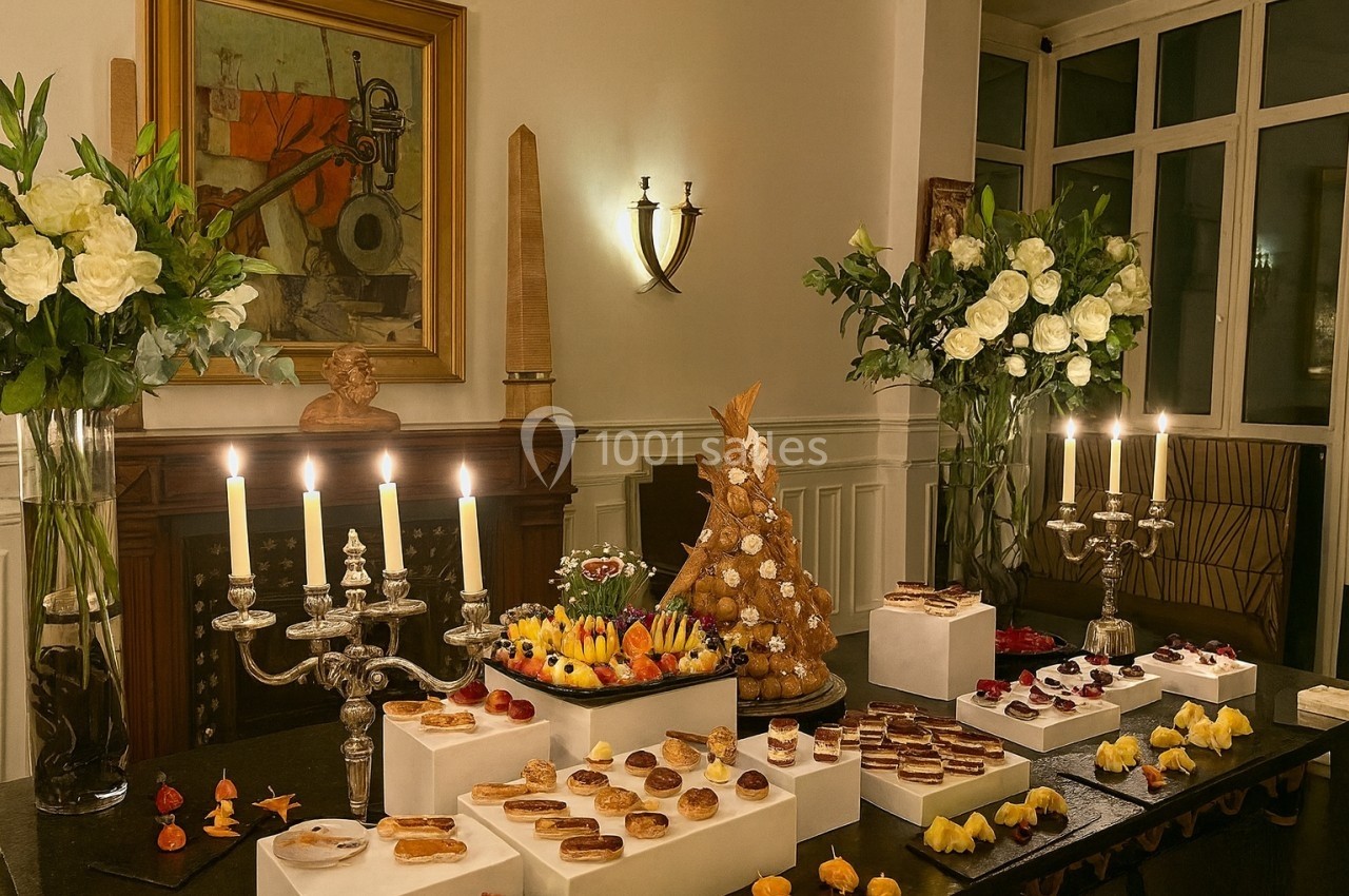 Table garnie de pâtisseries variées, éclairée par des chandeliers, avec des fleurs et une pièce montée en arrière-plan.