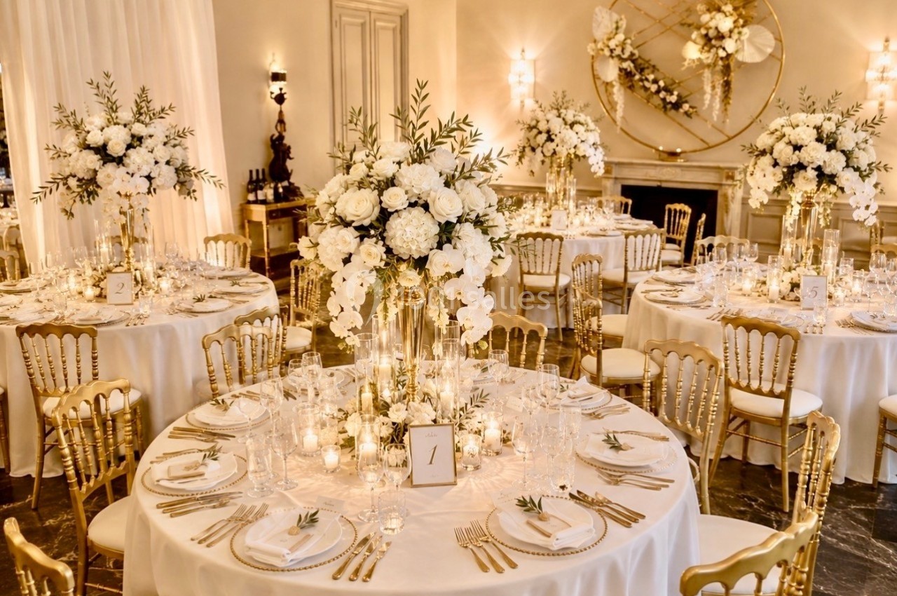 Salle de réception élégante avec tables rondes décorées de fleurs blanches, vaisselle raffinée et chaises dorées.
