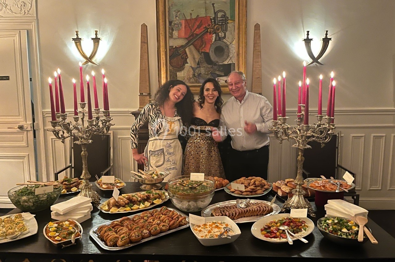 Trois personnes souriantes posent devant une table garnie de plats variés dans une salle élégamment décorée.