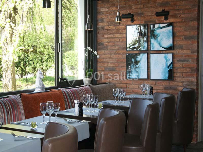 Salle de restaurant lumineuse avec tables dressées, chaises en cuir marron, mur en briques et grandes fenêtres donnant sur…