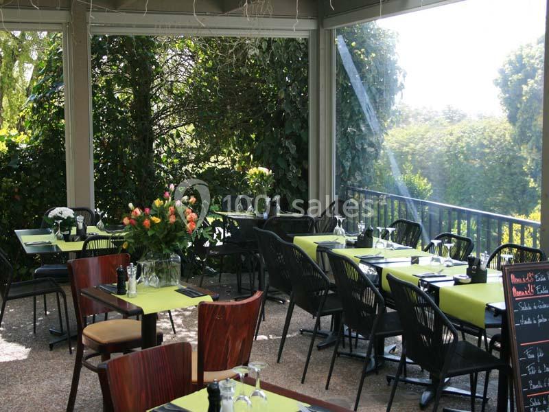 Terrasse d'un restaurant avec des tables dressées, nappes vertes, chaises noires et vue sur un jardin verdoyant.