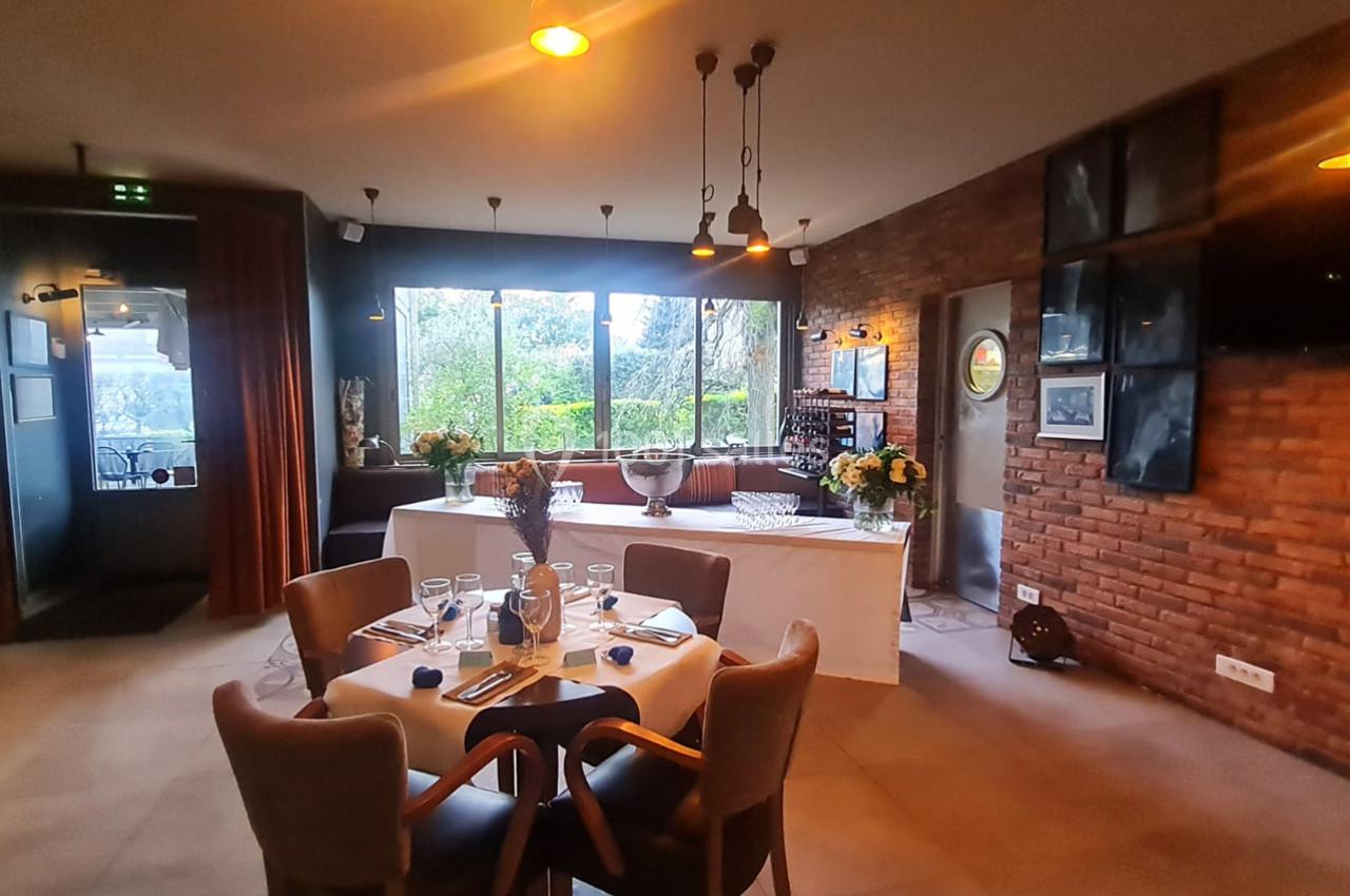 Salle de restaurant lumineuse avec table dressée, grandes fenêtres donnant sur un jardin et murs en briques.