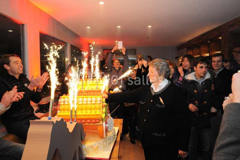 Une femme allume des cierges magiques sur un gâteau de fête entourée de personnes dans une salle éclairée.