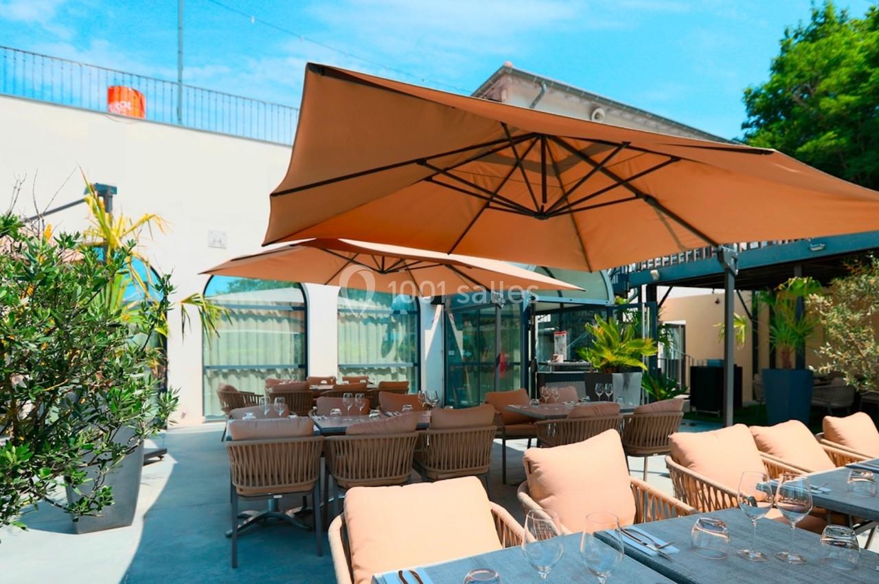 Terrasse extérieure avec tables dressées, chaises en rotin et grands parasols beige sous un ciel bleu.