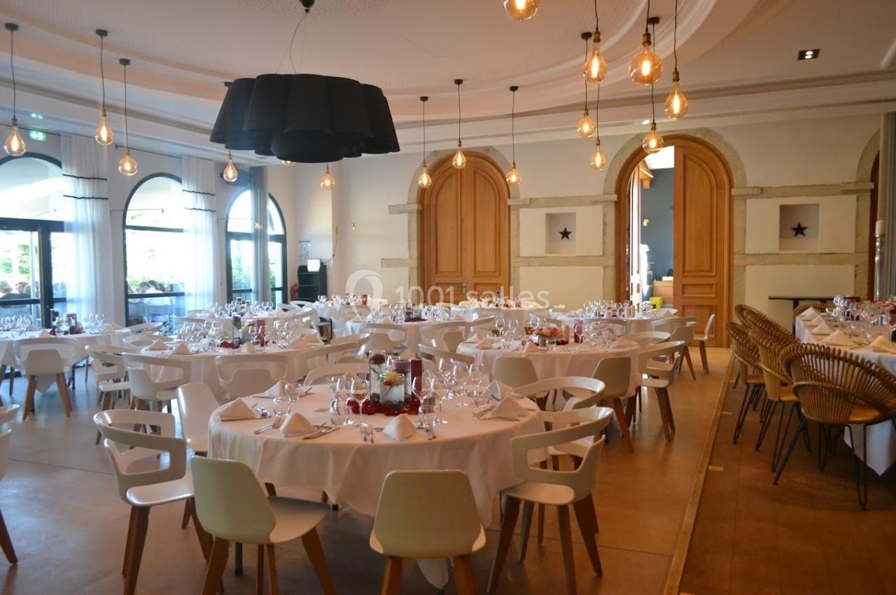 Salle de réception élégante avec tables rondes dressées, chaises blanches et luminaires suspendus.