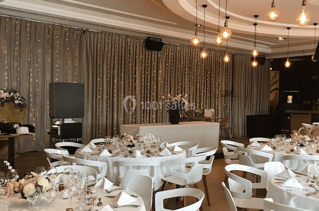 Salle de réception élégante avec tables dressées, chaises blanches et éclairage suspendu dans une ambiance chaleureuse.