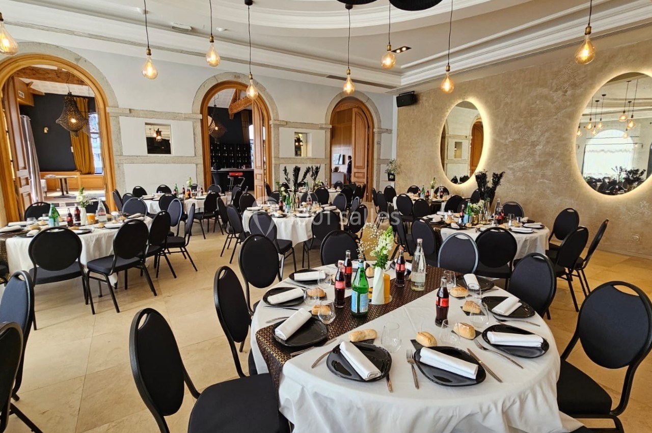 Salle de réception élégante avec tables rondes dressées, chaises noires et éclairage chaleureux.