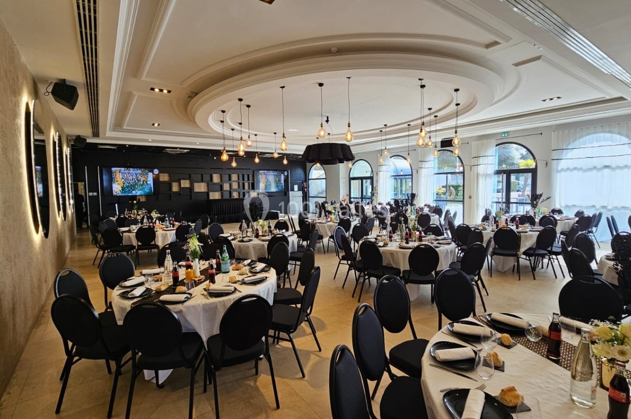 Salle de réception élégante avec tables rondes dressées, chaises noires et grandes baies vitrées laissant entrer la lumière…