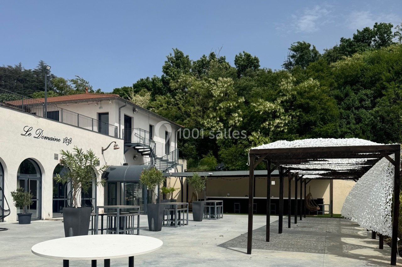 Terrasse extérieure avec tables, pergola moderne et bâtiment blanc entouré de verdure sous un ciel dégagé.