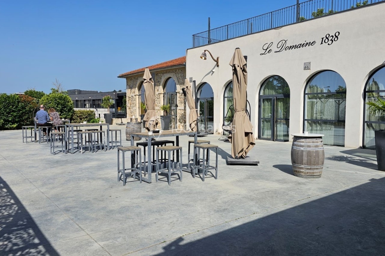 Terrasse ensoleillée avec tables hautes, parasols fermés et façade d'un bâtiment portant l'inscription ’Le Domaine 1838’.