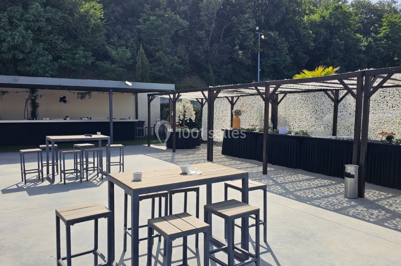 Terrasse extérieure aménagée avec tables hautes, chaises et pergolas, entourée de verdure en arrière-plan.