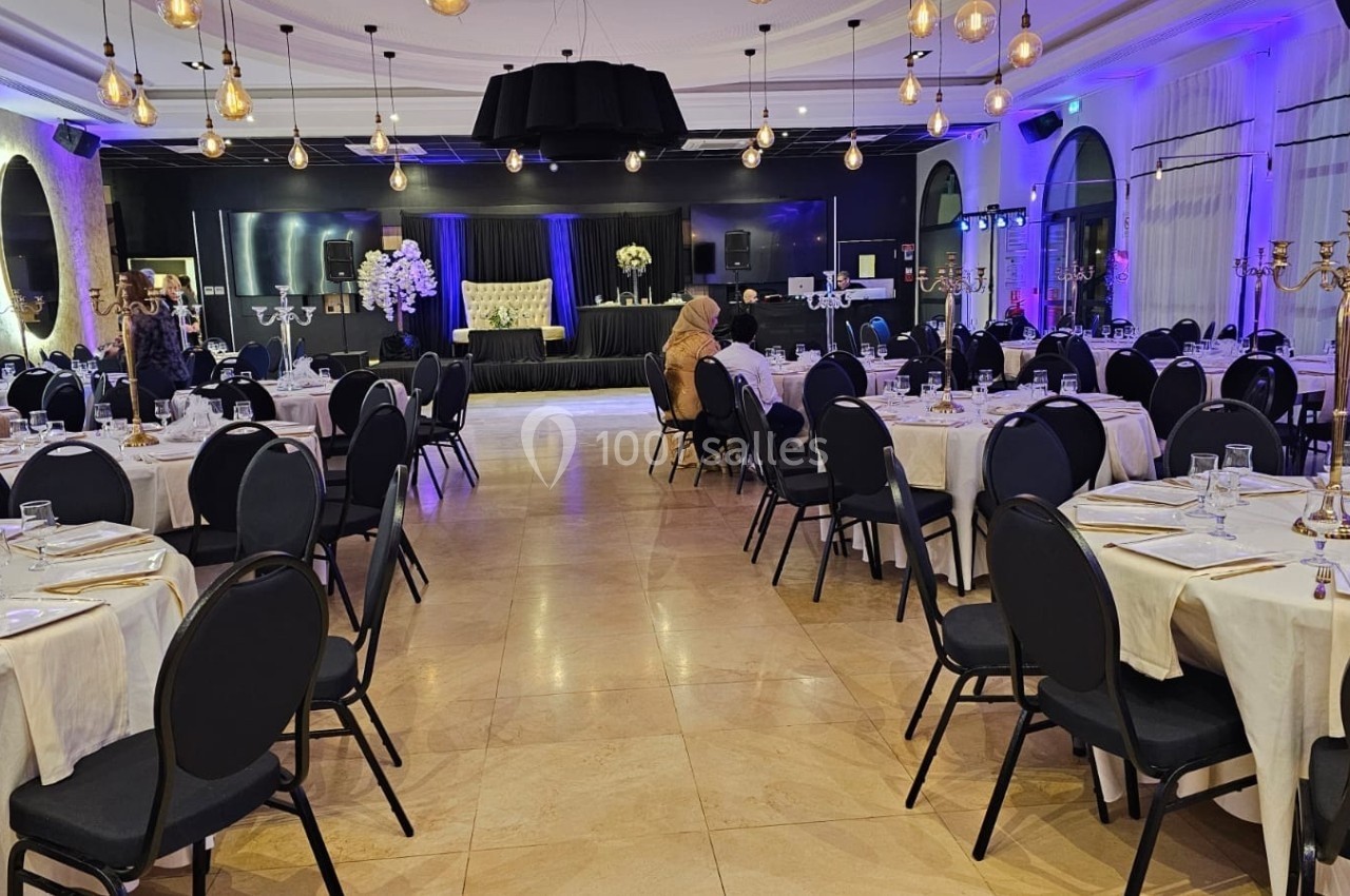 Salle de réception élégante avec tables dressées, chaises noires, éclairage tamisé et scène décorée en arrière-plan.