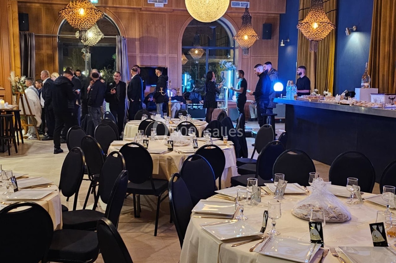 Salle de réception élégante avec tables dressées, chaises noires et invités debout près d'un bar éclairé.