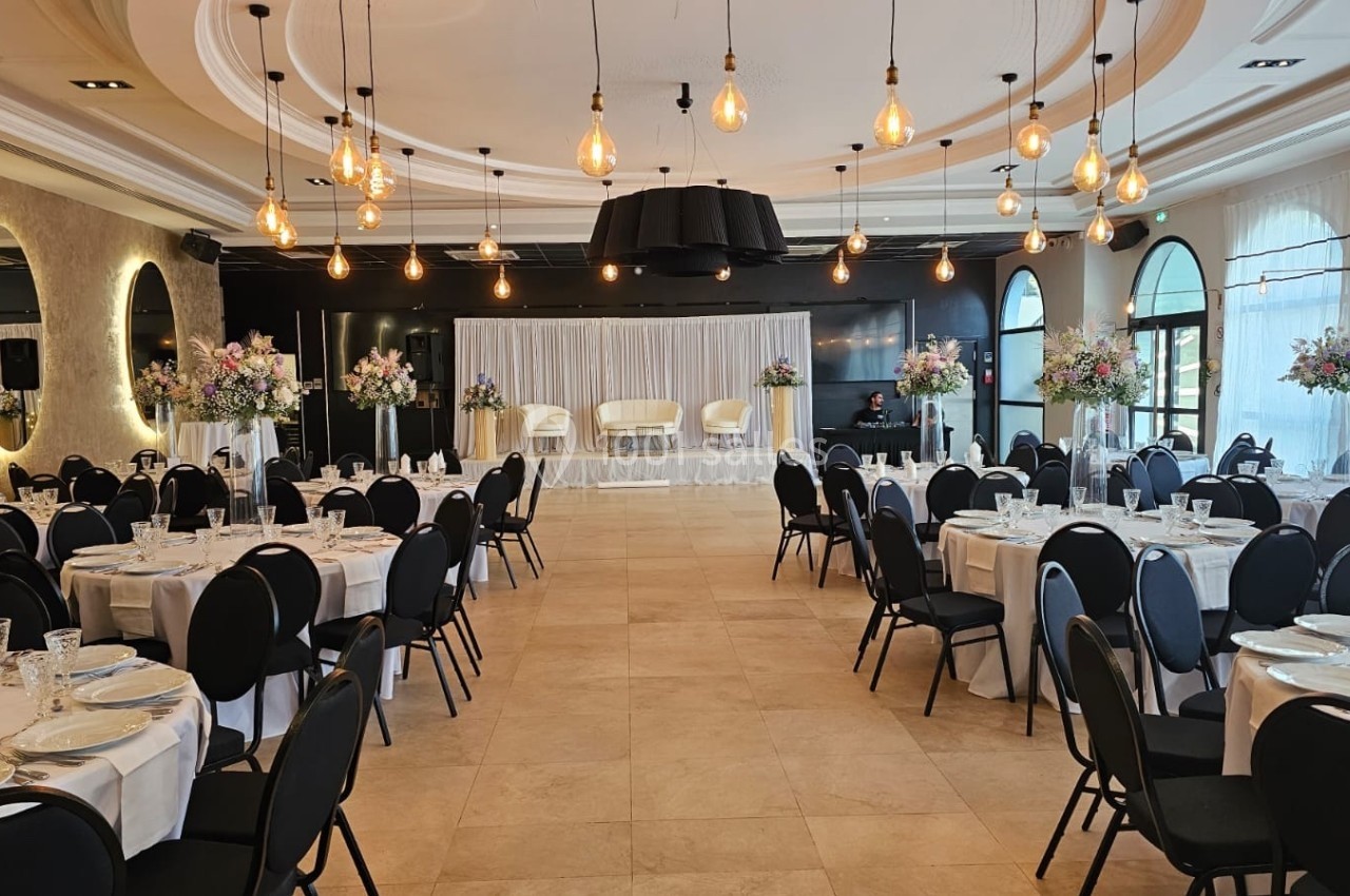 Salle de réception élégante avec tables dressées, chaises noires, décorations florales et scène en arrière-plan.