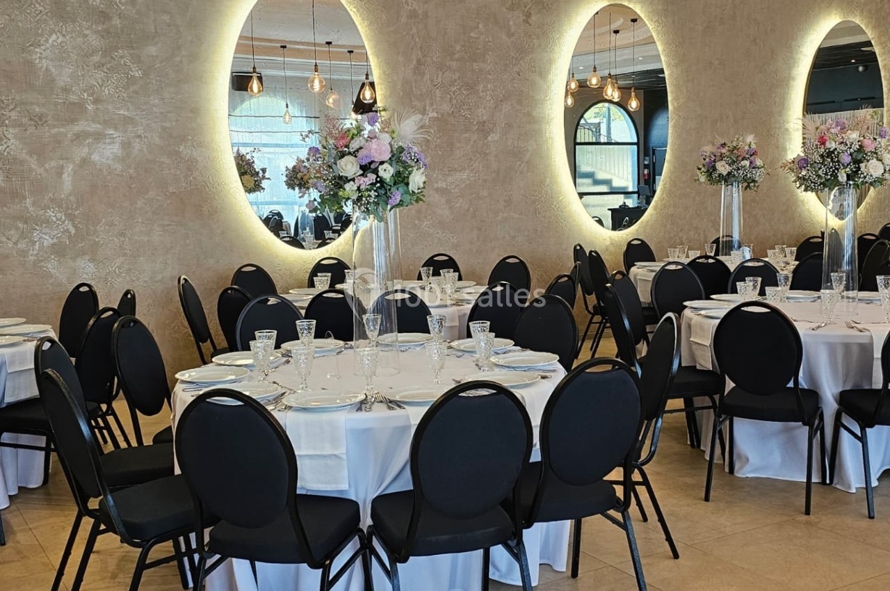 Salle de réception élégante avec tables rondes dressées, chaises noires et décorations florales sur fond de miroirs lumineux.